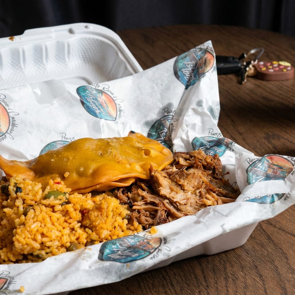 Takeout tray with yellow rice, pulled pork, cheese-topped fried plantain, dipping sauces, and a creamy beverage on a wooden table.
